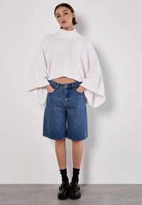 Weißer gestrickter, taillierter Pullover mit breiten Ärmeln, kombiniert mit blauen Denim-Bermuda-Shorts. Schwarze Schuhe und dunkle Socken vervollständigen das Outfit.