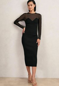 Femeie îmbrăcată într-o rochie midi neagră ajustat�ă, cu mâneci lungi din plasă și decolteu în formă de inimă, asociată cu sandale bej cu barete.