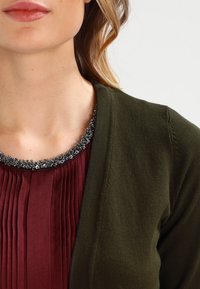 Oliwkowy kardigan z teksturowanym splotem, noszony na plisie w kolorze bordowym, z ozdobnym dekoltem wykończonym koralikami w srebrnych odcieniach.