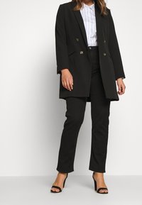 Sort skræddersyet blazer med dobbeltradet design og gyldne hardware-detaljer. Kombineret med sorte bukser og sorte stropper på højhælede sko.