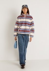 Φλις μπλούζα με κόκκινα και μπλε γεωμετρικά σχέδια, ψηλό γιακά, φορεμένη με μπεζ τζιν με άνετη εφαρμογή και τσάντα ώμου ανοιχτού μπλε χρώματος.