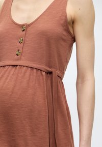 Αμάνικο φόρεμα σε σκουριά χρώμα με τρία καφέ κουμπιά και δεμένη ζώνη στη μέση σε χέρι και ώμο με ανοιχτό τόνο δέρματος.