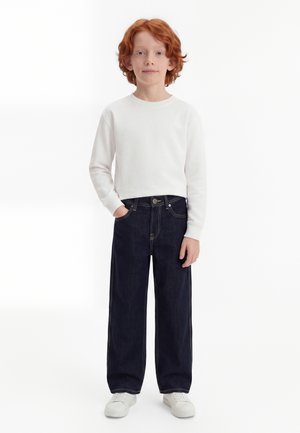 Kind mit roten Haaren, trägt ein weißes Langarmshirt, dunkle Jeans mit weitem Bein, weiße Sneakers, steht mit einer Hand in der Tasche.