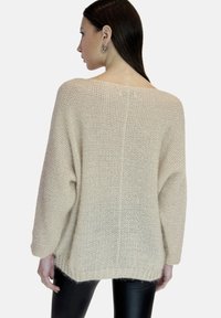 Beżowy sweter dzianinowy o luźnym kroju, z szerokimi rękawami i ribbowym wykończeniem. Prezentuje fakturowany materiał oraz otwarty tył.