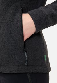 Donkergrijze fleece jack met een ritszak aan de linkerkant. Heeft een gladde textuur en subtiele stiksels. Groene merketiket zichtbaar.