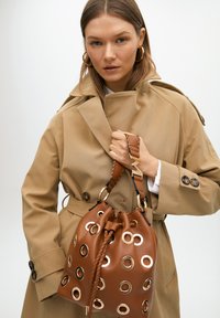 Brown leather bucket bag with metal grommets, braided handle, and gold accents. Worn by a model in a beige trench coat.