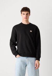 Zwarte sweatshirt met een ronde halslijn, geribbelde mouwen en zoom. Heeft een kleine CK-logo patch op de borst. Draagt met lichtblauwe jeans.