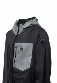 Giacca leggera nera e grigia con zip frontale, cappuccio, tasca sul petto con zip e patch con logo sulla manica. Texture liscia, design moderno.