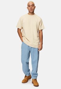 Beige Baumwoll-T-Shirt mit Rundhalsausschnitt, kombiniert mit hellblauen Jeans und braunen Schnürstiefeln. Einfaches Design, lockere Passform und einfarbige Farben.