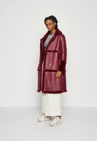 Bordeaux faux fur frakke med en notch-krave, forlommer og en afslappet pasform, sammen med lyse cargo-bukser og sølvfarvede sneakers.