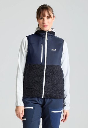Una mujer luce un chaleco azul marino y negro con cremallera y forro polar sobre una prenda blanca de manga larga, combinado con pantalones azul marino a juego, sobre un fondo gris liso.