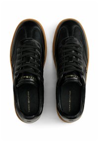 Paar schwarze Sneakers mit Lederbändern, braunen Sohlen sowie "Tommy Hilfiger"-Markenaufdruck auf Innensohlen und Zungen auf weißem Hintergrund.