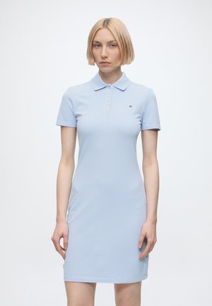Donna con capelli biondi corti che indossa un vestito polo azzurro a maniche corte, in piedi davanti a uno sfondo chiaro e semplice.