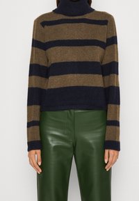 Vrouw in een bruin en marine gestreepte coltrui, gecombineerd met hoge groene leren broeken, staand tegen een effen achtergrond.
