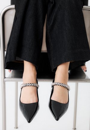 Zapatos de charol negros de punta con una brillante correa de rhinestones en el tobillo, combinados con pantalones de mezclilla oscuros.