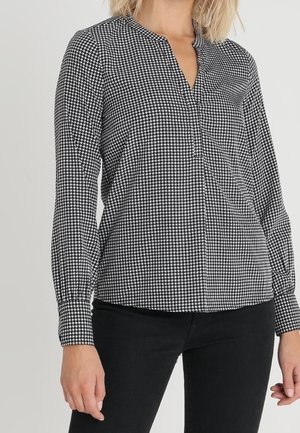 Kvinde iført en langærmet sort og hvid bluse med hundetandsmønster, kombineret med sorte bukser, stående foran en ensfarvet baggrund.