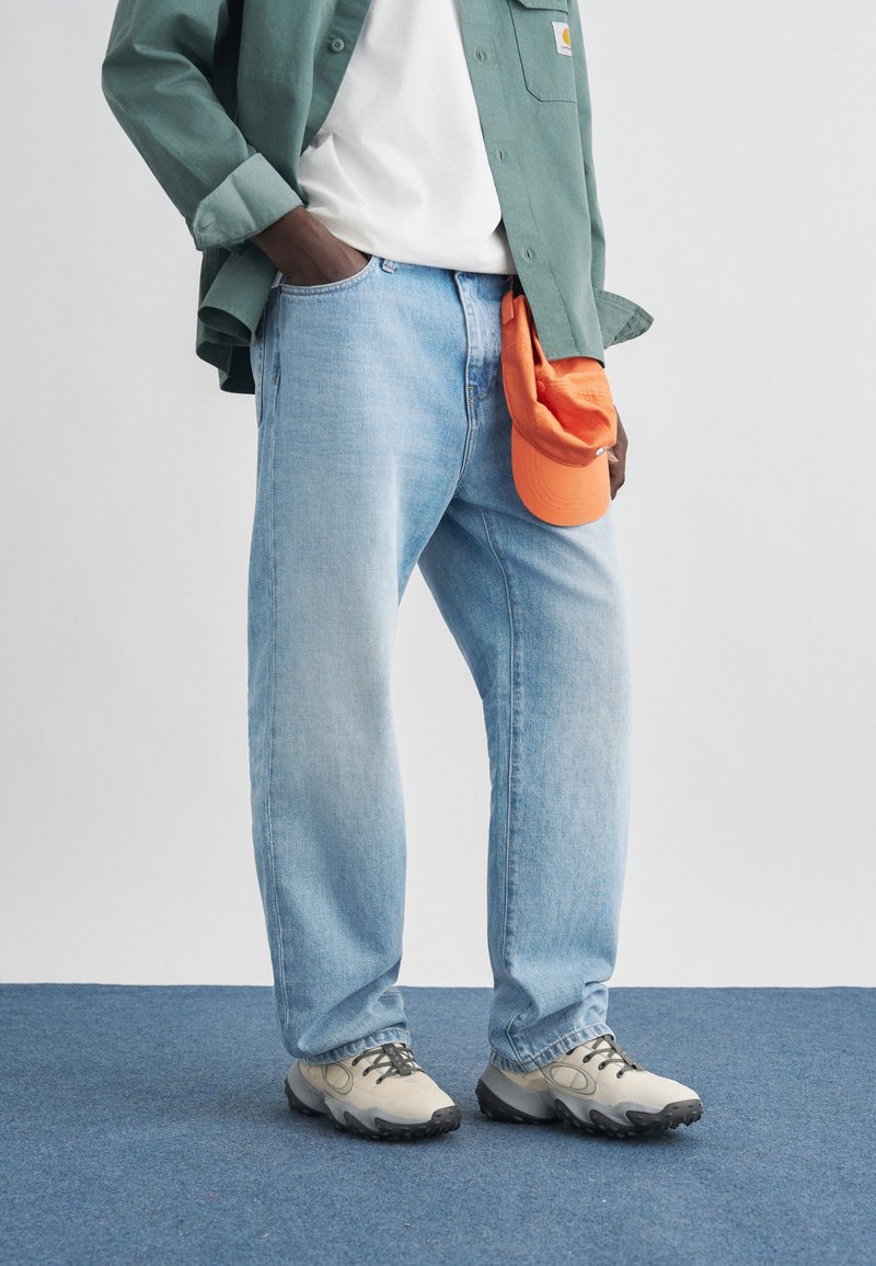 Person wearing light blue jeans, white sneakers, white shirt, and green jacket holding an orange cap against a plain background.