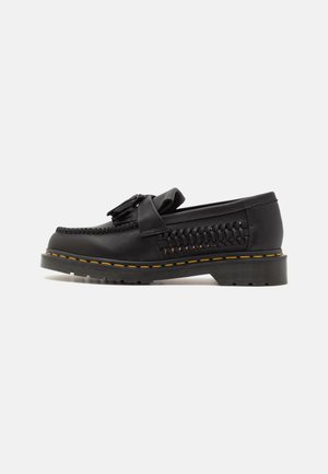 Mocassin en cuir noir avec côtés tissés, détail pompon sur le dessus, semelle épaisse à rainures, et surpiqûres jaunes autour du bord de la semelle.