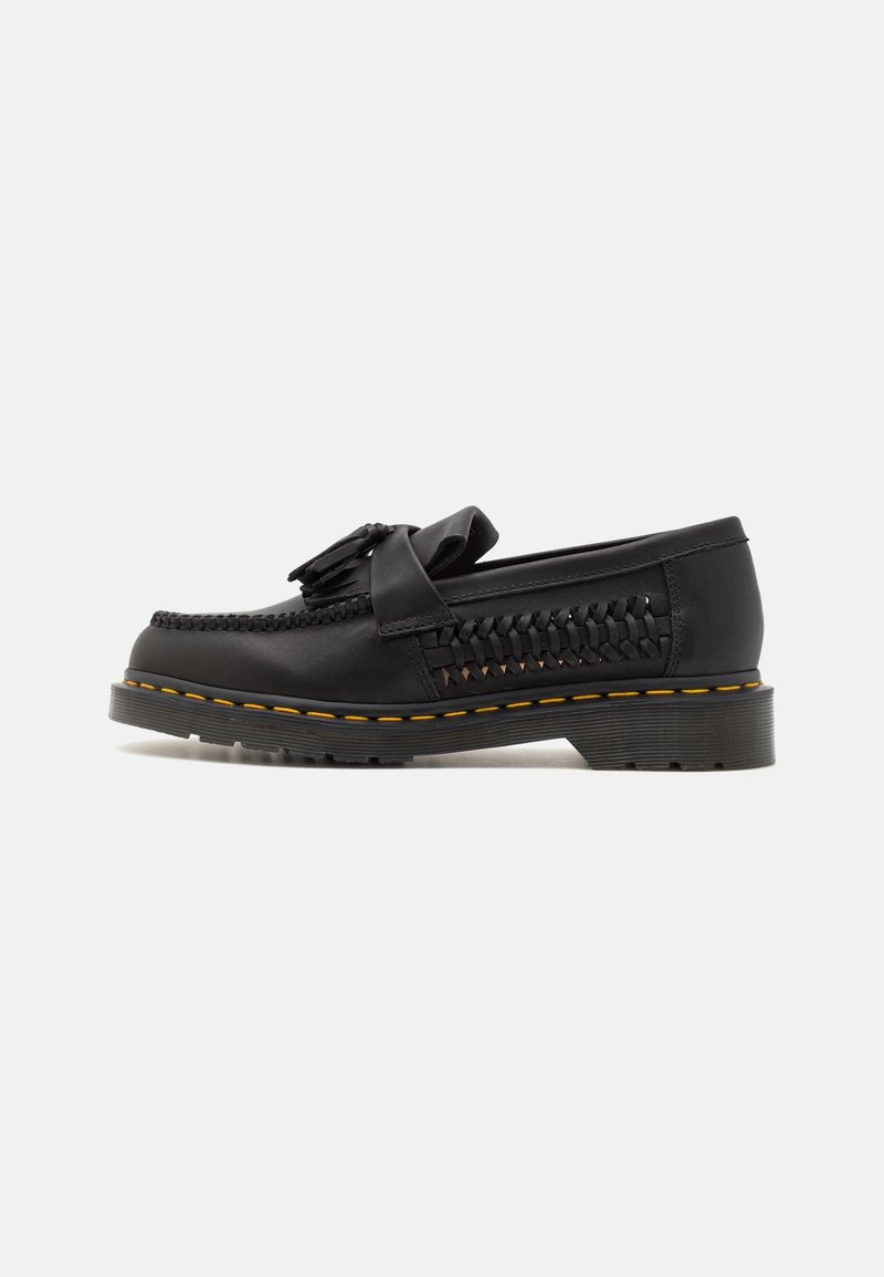 Mocassin en cuir noir avec côtés tissés, détail pompon sur le dessus, semelle épaisse à rainures, et surpiqûres jaunes autour du bord de la semelle.