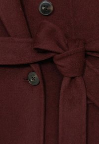 Bordeaux uldfrakke har et bundet stofbælte, en sort knap med fem huller og en blød tekstur, der fremhæver dens varme og struktur.