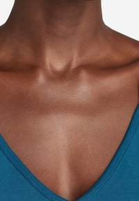 Teal V-paita, valmistettu sileästä ja istuvasta kankaasta. Läheltä otettu kuva korostaa ihon rakennetta ja olkaimen ääriviivoja vaaleaa taustaa vasten.