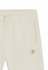 Crème kleurige sportieve broek met een trekkoord in de taille, zijzakken en een textuur met een hexagonaal patroon aan de zijkant. Geborduurd logo-detail.
