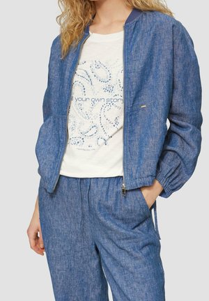 Nainen, joka pukeutunut siniseen denim-bomber-takkiin ja vastaaviin housuihin sekä valkoiseen graafiseen t-paitaan, jossa lukee "Kerro oma tarinasi."