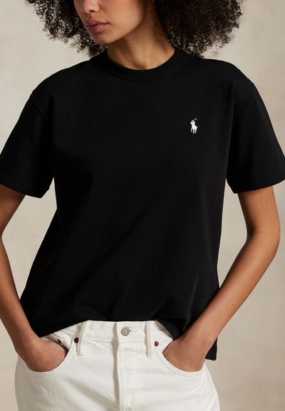 Nő fekete rövid ujjú pólóban, fehér hímzett logóval és fehér farmerral, zsebében a kezével, részben látható göndör hajjal.