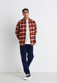 Camisa de cuadros para hombre en rojo y blanco con diseño de botones, combinada con pantalones azul marino y zapatillas negras con suelas blancas.