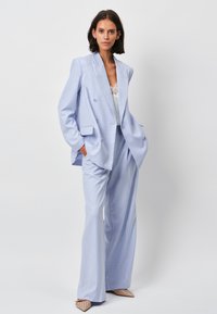 Lichtblauwe dubbelgeknoopte blazer met brede revers en twee voorkeurszakken; gecombineerd met bijpassende op maat gemaakte broek en nude studschoenen.