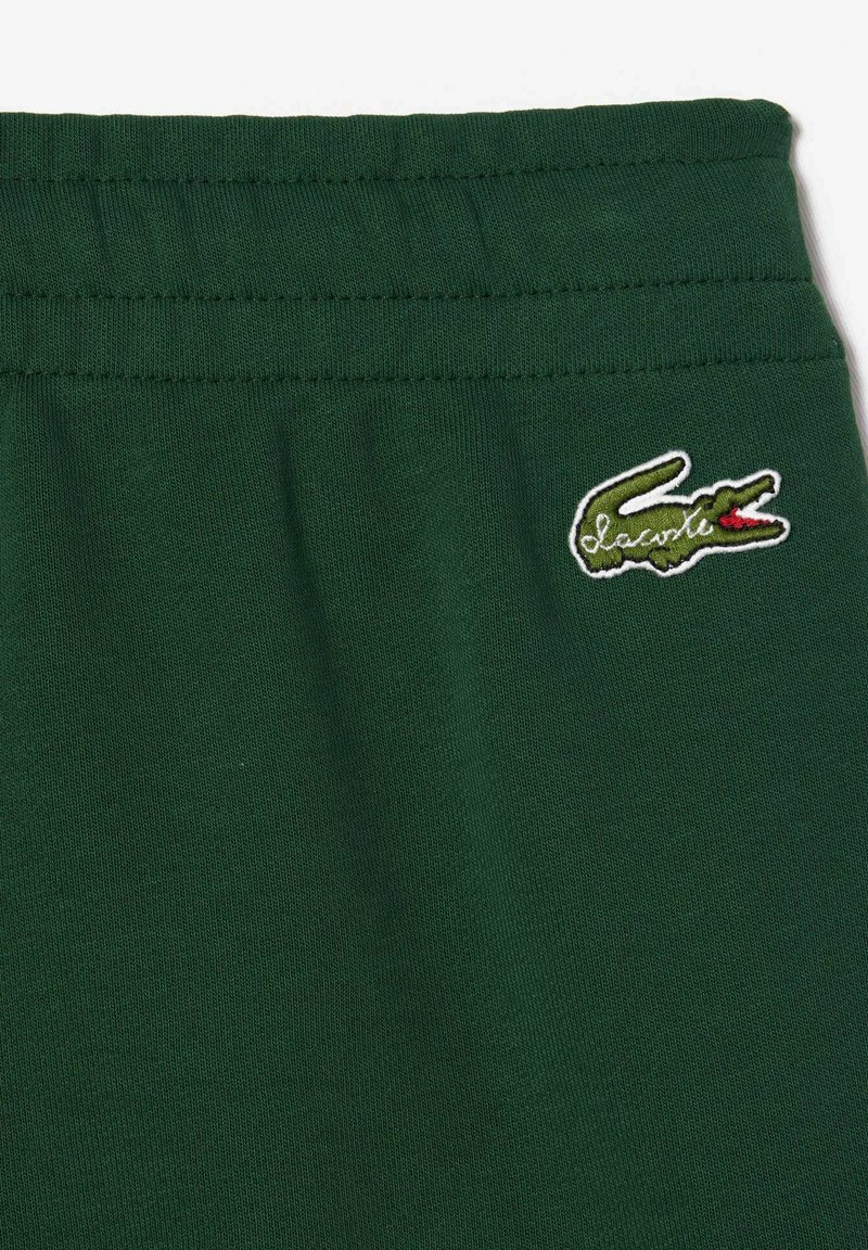 Zielone bawełniane spodnie dresowe z rypsowym paskiem w talii oraz małym haftowanym logo krokodyla w białym i czerwonym kolorze na lewym udzie.