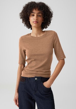 Frau mit lockigem Haar, die ein braunes kurzärmeliges Oberteil und dunkelblaue Jeans trägt, steht mit einer Hand in der Tasche vor einem einfarbigen Hintergrund.