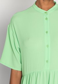 Mintunvihreä napitettava mekko, jossa on mandariinikaulus, lyhyet hihat ja teksturoitu kangas. Mekossa on kerrostettu hame ja napitettu yksityiskohtia.