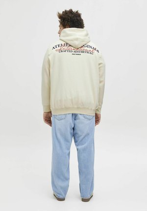 Beige hættebluse med snøre, der har tekst på ryggen i kontrastfarver. Løs pasform, parret med lyseblå baggy jeans.