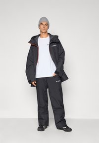 Chaqueta negra impermeable con detalles en gris, cremallera naranja y pantalones negros a juego. El modelo lleva una camiseta blanca y un gorro gris.
