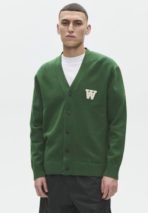 Jonge man met kort haar, gekleed in een groen vest met knopen en een witte "W" patch, en zwarte broek, staand tegen een effen achtergrond.