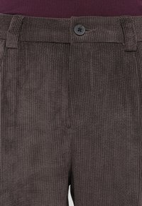 Gros plan de pantalon en velours côtelé marron foncé côtelé à l'avant avec un bouton noir et des passants de ceinture, porté avec un haut en tricot bordeaux foncé.