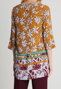 Tunique florale en jaune moutarde avec des fleurs blanches, présentant un design à motifs mixtes sur l'ourlet, des manches trois-quarts et une coupe décontractée.