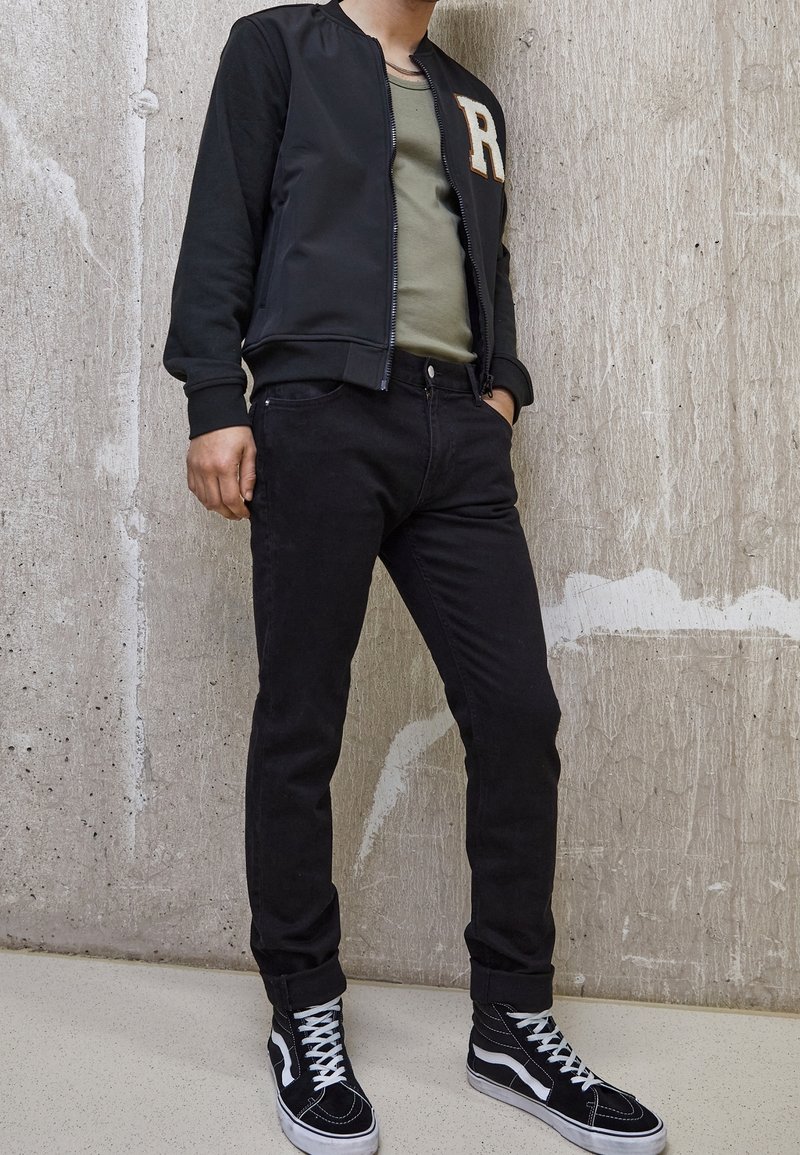 Chaqueta bomber negra con bordado de la 'R', camiseta verde oliva, jeans negros y zapatillas altas en blanco y negro. Fondo de hormigón texturizado.