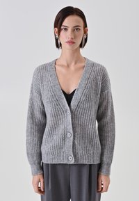 Cardigan lavorato a maglia grigio chiaro con scollo a V, dotato di cinque bottoni, texture a coste e maniche lunghe. Indossato sopra un top nero.
