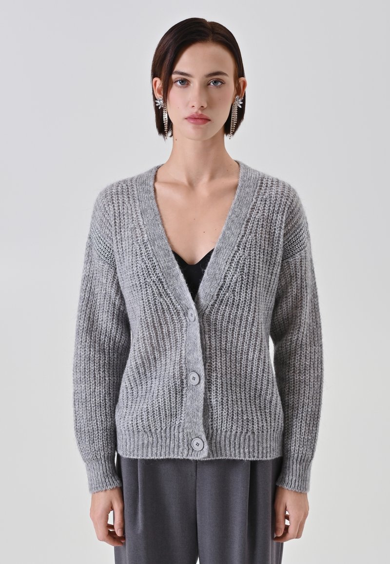 Cardigan lavorato a maglia grigio chiaro con scollo a V, dotato di cinque bottoni, texture a coste e maniche lunghe. Indossato sopra un top nero.
