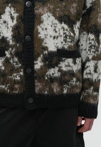 Pletena kardigan u smeđoj, krem i crnoj boji s pikastim uzorkom. Ima pet crnih dugmadi, rebraste manžete i dva otvorena prednja džepa.