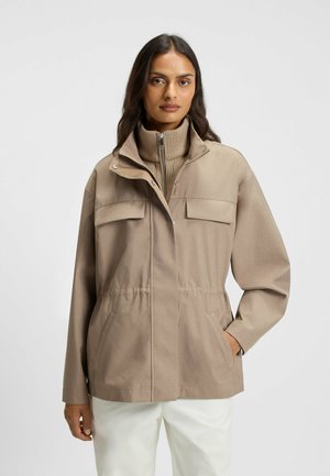 Beige takki, jossa on korkea kaulus, vetoketjusuljenta ja kaksi rintataskua. Varustettu joustavalla vyötäröllä ja sileällä, mattapintaisella rakenteella.