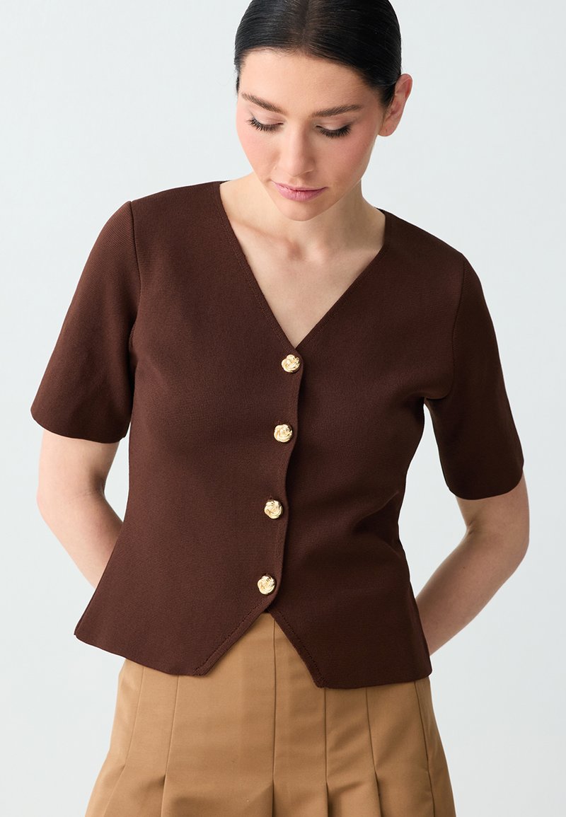 Femme portant un chemisier marron à manches courtes et col en V, orné de boutons dorés en forme de fleurs, assorti à un pantalon plissé beige, regardant vers le bas.
