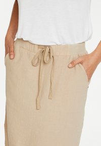 Gros plan d'une personne portant une jupe beige à cordon avec des poches, les mains reposant dans les poches, associée à un top blanc.