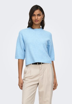 Kvinde iført lyseblå kortærmet sweater, beige højtaljede bukser og gyldne ringearreringe, stående mod en ensfarvet lys baggrund.