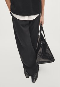 Pantaloni neri a gamba larga, orlo della camicia bianca visibile, scarpe nere con punta a punta, che tiene una borsa tote nera con texture. Design minimalista, look elegante.