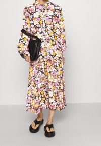 Robe maxi florale bleu foncé avec des fleurs vibrantes roses, jaunes et blanches. Manches longues, fermeture à boutons et jupe fluide. Associée à des sandales noires.