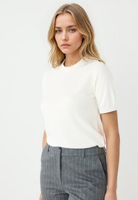 Weißes kurzärmliges Strickoberteil mit rundem Halsausschnitt, kombiniert mit grauen Nadelstreifen-Hosen. Weiche Textur und taillierter Schnitt.