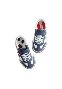Fehér tornacipők tengerészkék díszítésekkel, tépőzáras pánttal, fűzőkkel és játékos figura kitűzővel. Texturált talp és színes bélés részletek.